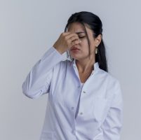 tired young female doctor wearing medical robe holding nose with closed eyes isolated on white background with copy space